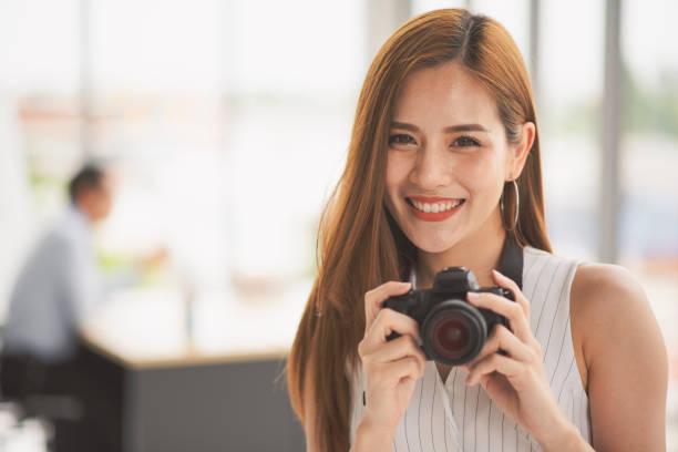 istock Successful business young Asian beautiful woman looking at camera and hold a camera while colleague discuss or meeting behind her in the office 1131617859