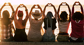 Love and meditation lifestyle concept - group of women looking at the sunset on the ocean and make the hearth with hands - romantic golden light and valentine's day concept - friendship forever