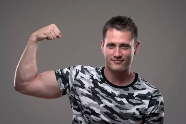 Portrait of athletic muscular fitness male model in army shirt flexing arm and showing bicep muscles.