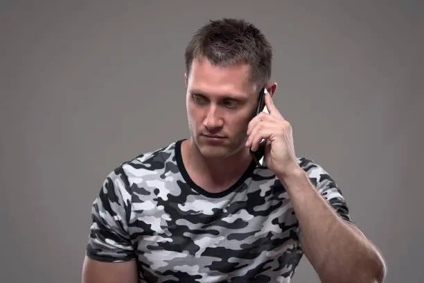 Serious disappointed young man talking on the mobile phone and looking down on gray background.