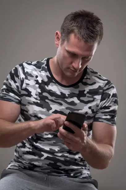 Young fit handsome adult man using cellphone typing messages or surfing the net over gray background.