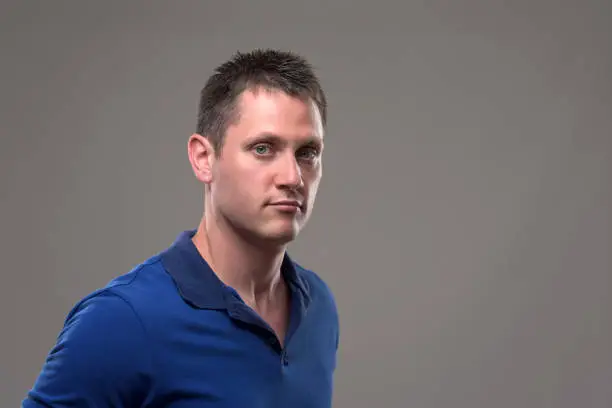 Moody portrait of serious confident young man in blue polo shirt looking at camera on gray background.