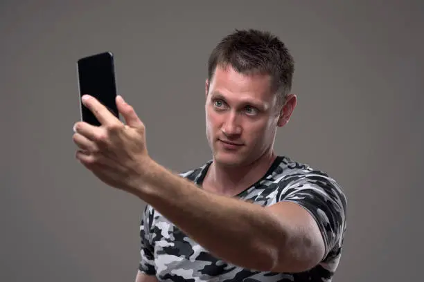 Handsome young man in camouflage pattern t-shirt taking photos with mobile phone on gray background.