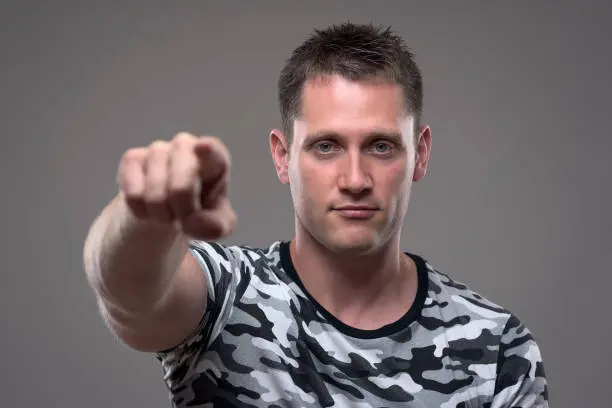 Portrait of confident successful young athletic man in army shirt pointing finger at camera choosing you.