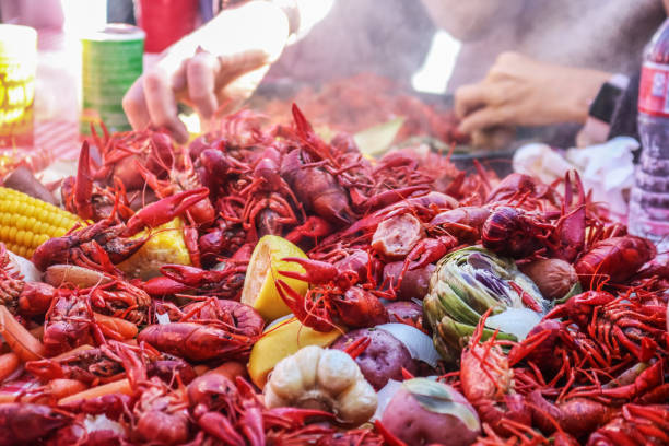Boiled crawfish and vegetables piled on red checked tablecloth with eating tray and arm of person eating bokeh behind - shallow focus stock photo