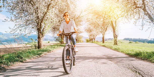 feliz mujer sonriente monta una bicicleta en la carretera del campo bajo árboles de flor. la primavera se acerca a la imagen conceptual. - comming fotografías e imágenes de stock