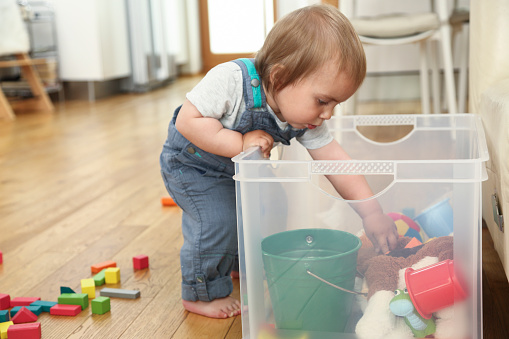 Little boy standing by plastic box with toys and looking in it's content. Trying to reach his toys.