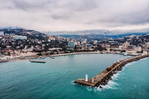 Aerial view of Yalta embankment from drone, old Lighthouse on pier, sea coast landscape and city buildings on mountains, beautiful winter panorama of European resort, Crimea.