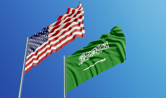 American and Saudi Arabian flags are waving with wind over  blue sky. Low angle view. Dispute and conflict concept. Horizontal composition with copy space.