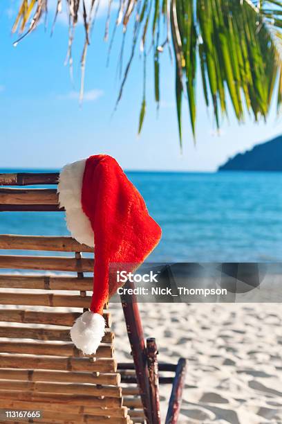 Santas Red Hat Colgar En Una Silla De Playa Bajo Una Palmera Foto de stock y más banco de imágenes de Aire libre