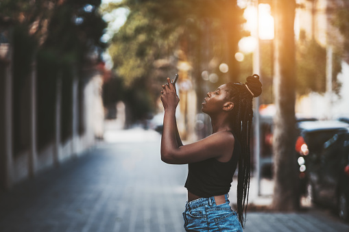 A charming African teen with braided hair is using the camera of her smartphone to photoshoot something on the street, above; shallow depth of field, strong bokeh, sunny day in Barcelona, Spain