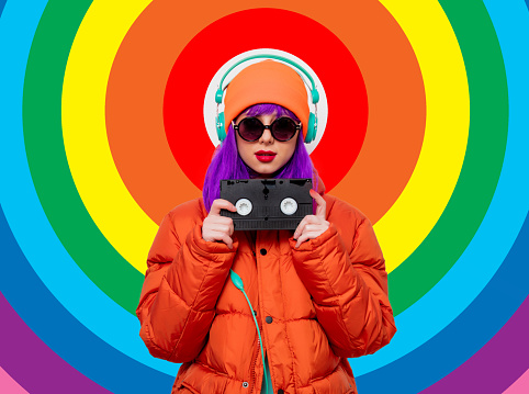 Portrait of a beautiful girl with purple hair in orange hat and jacket and with headphones and VHS cassette on rainbow background. Trendy style