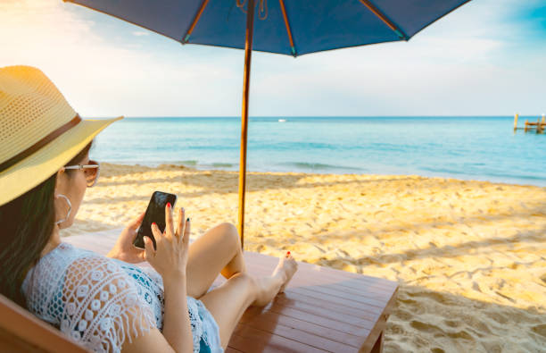 asiatische frau mit hut sitzt am sandstrand unter sonnenschirm und nutzt im sommerurlaub mit smartphone. mädchen im lässigen stil entspannen und genießen sie urlaub am tropischen paradies strand. sommervibes. - sea summer umbrella beach stock-fotos und bilder