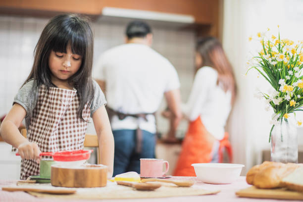 a família cozinha. a filha prepara o pó da massa. pai e mãe estão atrás e cozinhando. todos têm aventais. viva em casa na cozinha. - family with one child domestic life caucasian love - fotografias e filmes do acervo