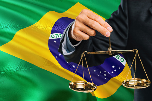 Brazilian Judge is holding golden scales of justice with Brazil waving flag background. Equality theme and legal concept.