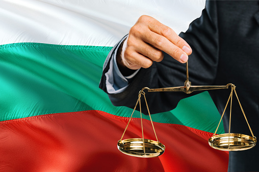 Bulgarian Judge is holding golden scales of justice with Bulgaria waving flag background. Equality theme and legal concept.