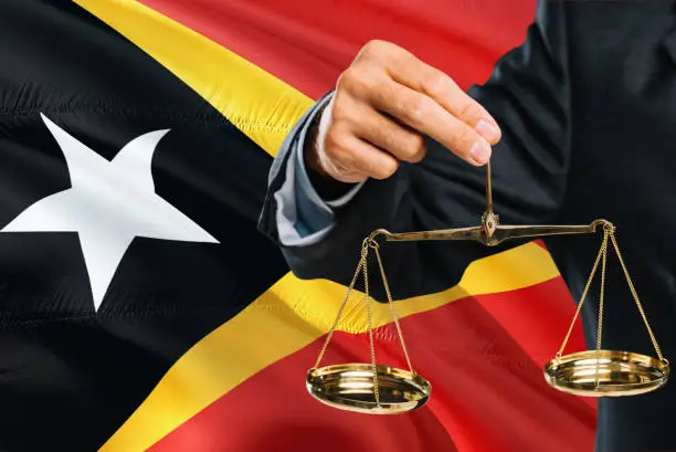 Photo of Timorese Judge is holding golden scales of justice with East Timor waving flag background. Equality theme and legal concept.