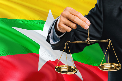 Burmese Judge is holding golden scales of justice with Myanmar waving flag background. Equality theme and legal concept.
