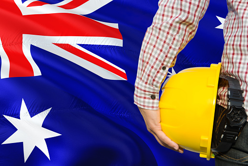 Australian Engineer is holding yellow safety helmet with waving Australia flag background. Construction and building concept.