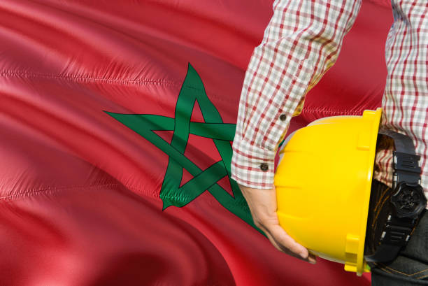 Moroccan Engineer is holding yellow safety helmet with waving Morocco flag background. Construction and building concept.