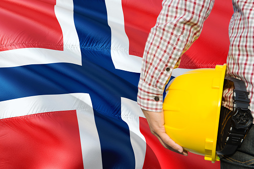 Norwegian Engineer is holding yellow safety helmet with waving Norway flag background. Construction and building concept.
