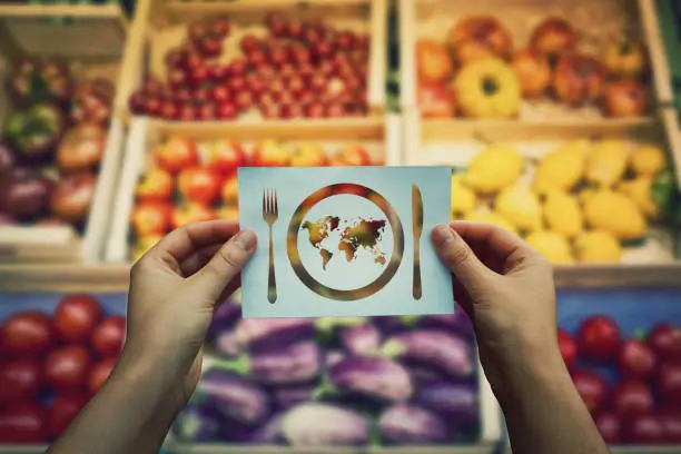 Global hunger issue, water supply problem. Hands holding a paper sheet with world map in a plate with knife and fork icon over market shelves background. International craving and starvation metaphor.