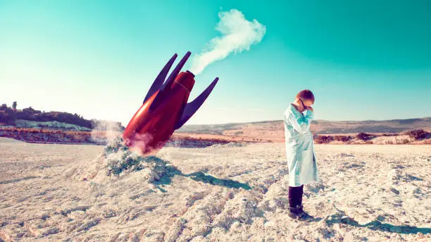 Child around 10 years old stands on a field after a failed rocket launch. He covers his head in his hands. He wears a lab coats like a real scientist.
