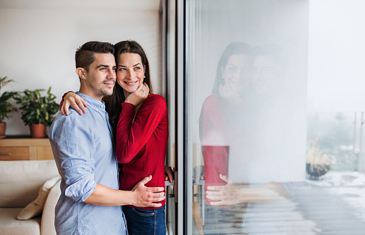 Young happy couple in love standing by the window at home, hugging. Copy space.
