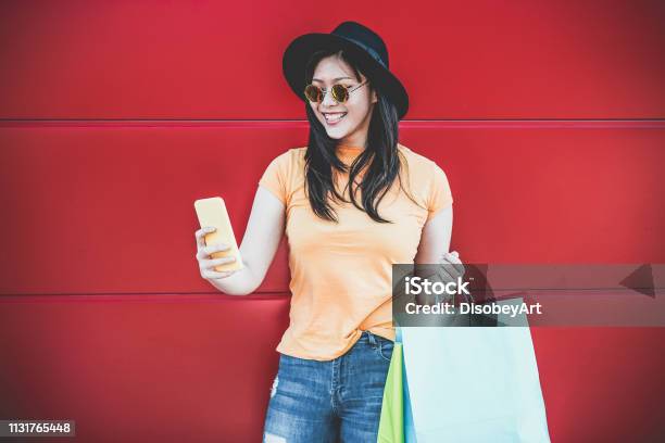 Happy Trendy Asian Girl Using Shopping App Holding Bags Outdoor Young Woman Having Fun Buying New Clothes On Web With Smartphone Retail Tech Sales And Buyer Addiction Concept Focus On Face Stock Photo - Download Image Now
