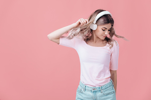 beautiful blonde woman in pink t-shirt and white headphones listens to music and smiling with closed eyes on pink background