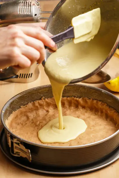 Two female caucasian hands pour cheesecake cream into a black baking pan, baking utensils in the background - side view, portrait orientation