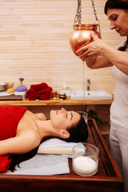 istock Dark-haired master in white uniform pouring stream of oil on client face 1131793102