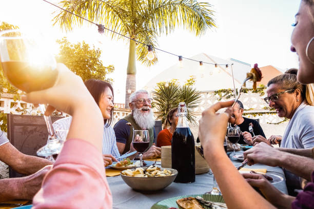日没時間中に夕食をやって幸せな家族-屋外で一緒に楽しい食事を持つ多様な友人のグループ-ライフスタイルの人々、食品や週末の活動の概念 - wine culture ストックフォトと画像