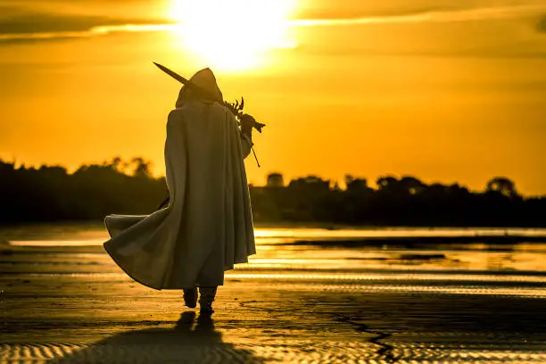 Portrait of assassin in white costume with the sword at the sea. Sunset time. Beautiful orange colors. Silhouette of man.