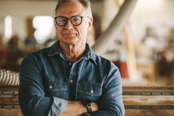 Proud carpentry workshop owner Portrait of successful senior man in eyeglasses standing in his carpentry workshop. Proud carpentry workshop owner standing with his arms crossed. carpenter portrait stock pictures, royalty-free photos & images