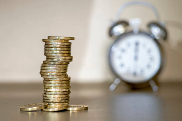 istock Yellow shiny coins stacked in a pile and the blurry clock on the differentiated light shadow background. 1131821765