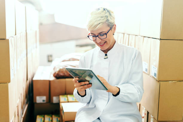 Happy Caucasian blonde employee suing tablet for work while sitting around boxes in food factory.