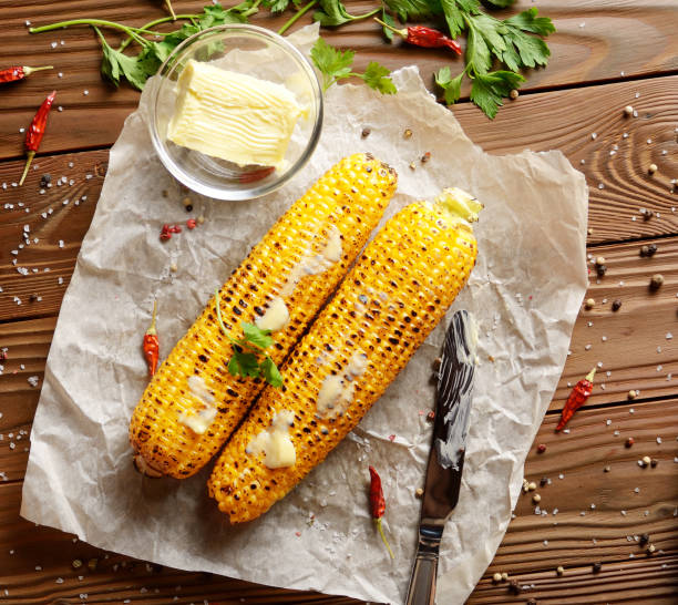 Top view of kitchen table with flat lay grilled sweet corn cob under melting butter and greens on baking paper
