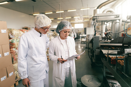Dos profesionales de calidad en uniformes estériles blancos que verifican la calidad de los palos de sal mientras están de pie en la fábrica de alimentos. photo