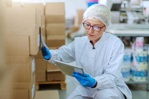 istock Caucasian blonde employee in sterile uniform crouching next to boxes and dealing with logistic of products. Food factory interior. In hand tablet. 1131839681
