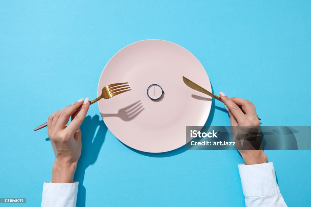 White plate with round whatch shows six o'clock served knife and fork in a girl's hands on a blue background. Time to eat and diet concept. Top view. Round watch of six o'clock and woman's hand with fork and knite in agirl's hands on a blue background with shadows. Time to lose weight, eating control or diet concept. Place for text. Fasting - Activity Stock Photo
