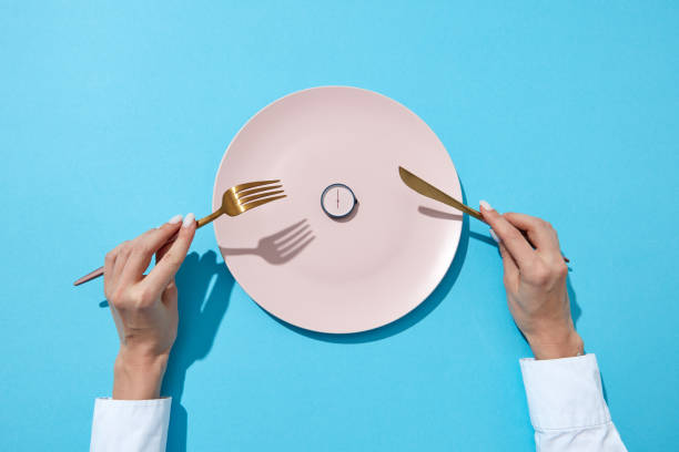 a placa branca com whatch redondo mostra que seis horas serviram a faca e a forquilha nas mãos de uma menina em um fundo azul. tempo para comer e conceito da dieta. vista superior. - prato vazio - fotografias e filmes do acervo