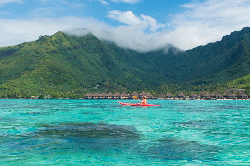 This boy is kayaking in the lagoon surrounding the island of Moorea, French Polynesia.  In the background are seen some of the mountain peaks in the middle of the island as well as some overwater bungalows jutting out into the lagoon.  The water surrounding the island is a very clear turquoise and aquamarine color.