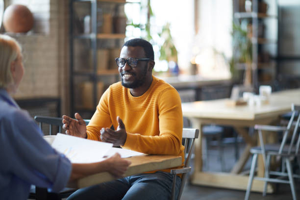 istock Smiling handsome young black man in bright sweater sitting at table and gesturing hands while presenting his idea to colleague 1131853788