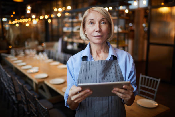istock Content confident attractive restaurant waitress in gray apron standing in modern banquet room and viewing order on tablet while looking at camera 1131854856