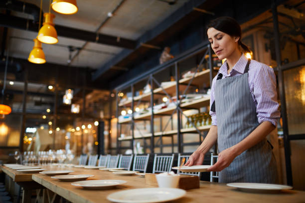 istock Serious young waitress in stripped apron putting plates on banquet table while serving long table alone in restaurant 1131854860