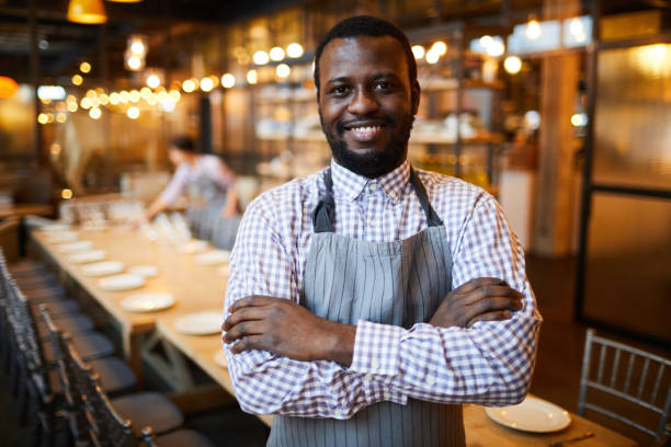 istock Cheerful handsome young black waiter with beard wearing gray stripped apron standing against long table and crossing arms on chest while looking at camera 1131854961