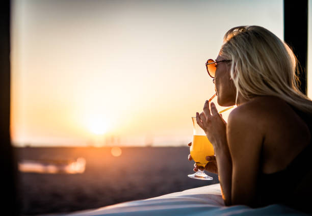 młoda kobieta pije letni koktajl na łóżku plażowym o zachodzie słońca. - juice drinking women drink zdjęcia i obrazy z banku zdjęć