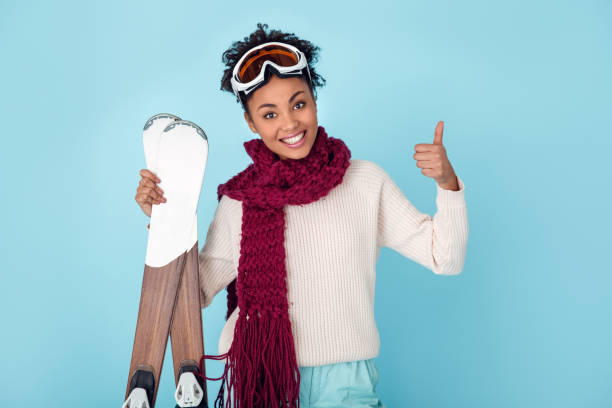 joven mujer africana aislado en azul pared estudio invierno deporte esquí pulgar hasta - snow gear fotografías e imágenes de stock