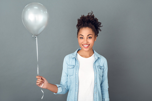 Young african female student isolated on grey wall standing holding grey balloon looking camera smiling happy